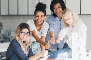 Fair-haired female it-specialist fooling around with friends, sitting at workplace and laughing. Excited japanese manager posing with smile, standing beside blonde colleague in office..